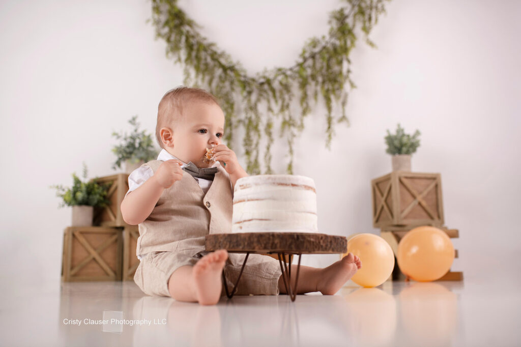 A toddler in a beige outfit sits on the floor eating cake from a small white cake in front of him. Behind him are two yellow balloons, green plants, and wooden crates with greenery hanging on the wall. Cristy Clauser Photography