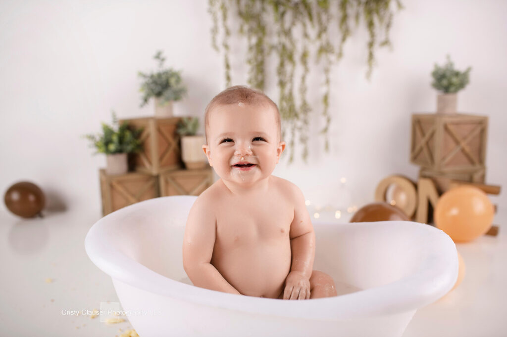 A smiling baby sits in a white bathtub, surrounded by brown and beige balloons, plants, and wooden decor. The background is softly decorated, creating a warm and cheerful atmosphere. Cristy Clauser Photography