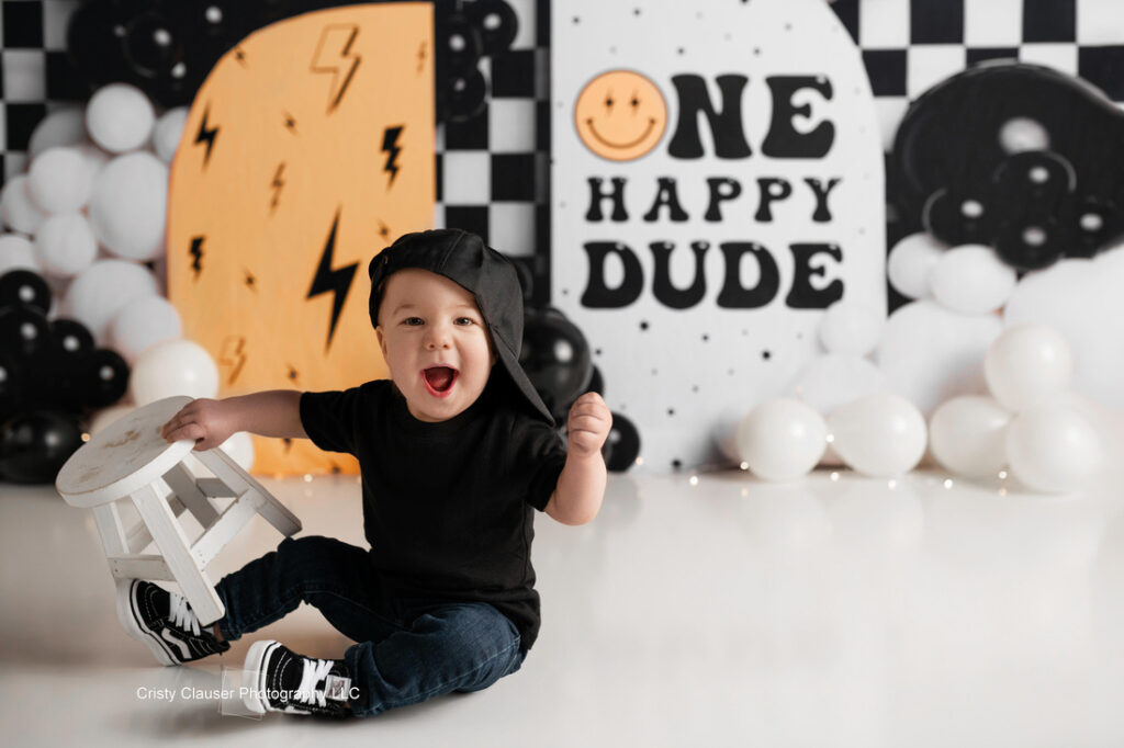 A smiling toddler in black clothes and a sideways cap sits on the floor holding a small white stool, with a “ONE HAPPY DUDE” birthday backdrop and black, white, and yellow balloons behind him. Cristy Clauser Photography