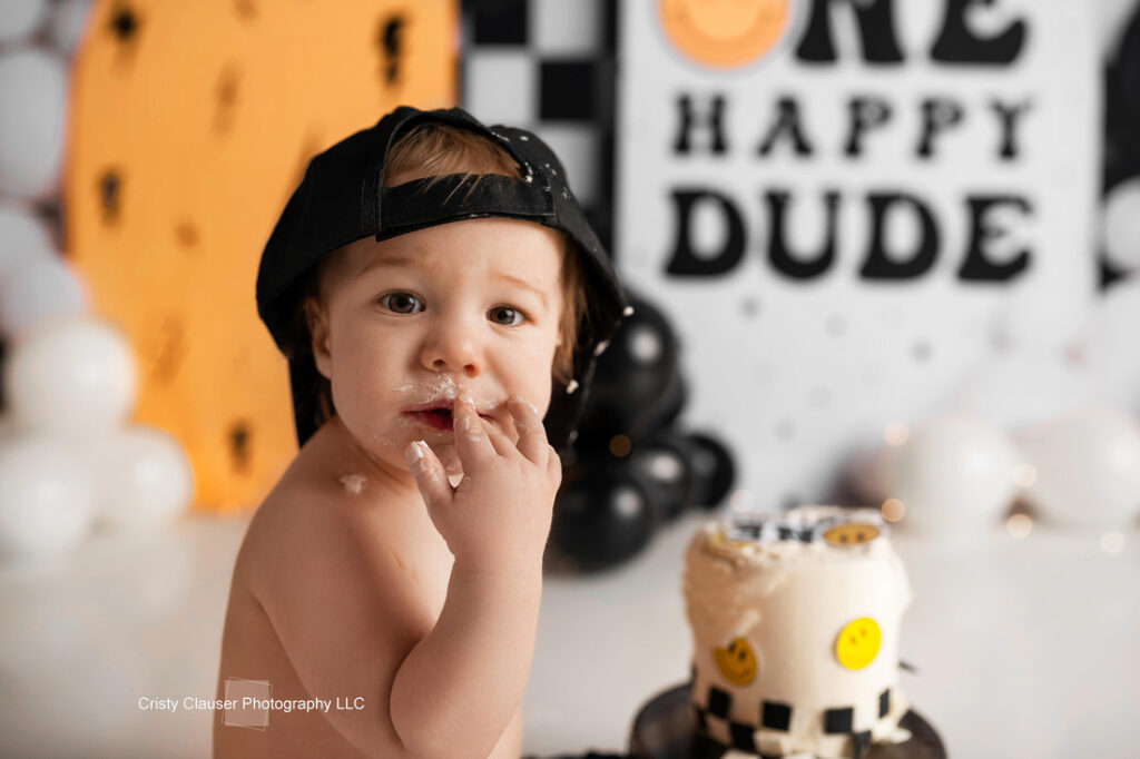 A toddler wearing a backwards black cap has cake on his face and fingers, standing in front of a small cake with smiley faces. The background features black and white balloons and a sign that reads "ONE HAPPY DUDE. Cristy Clauser Photography
