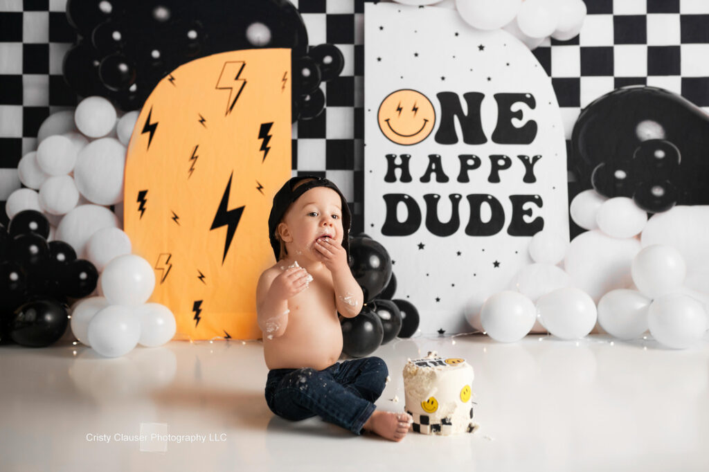 A baby wearing jeans and a black cap sits on the floor eating cake. Behind him, a black-and-white balloon display and a sign read "ONE HAPPY DUDE" in bold letters. The scene has a fun, celebratory atmosphere. Cristy Clauser Photography
