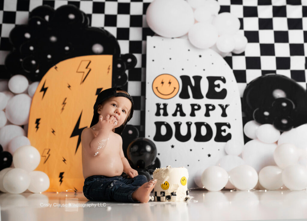 A toddler wearing a backward cap sits shirtless in front of a small cake with smiley faces, surrounded by black and white balloons and a backdrop that reads "ONE HAPPY DUDE. Cristy Clauser Photography