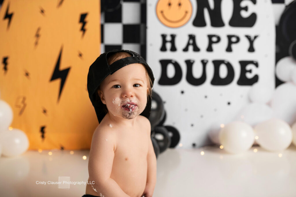 A toddler wearing a backward black cap and covered in cake sits in front of a “One Happy Dude” sign with a smiley face, surrounded by white and black balloons and festive decorations. Cristy Clauser Photography