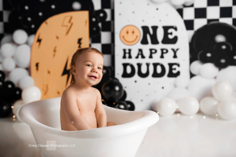 A smiling toddler sits in a white tub, surrounded by white, black, and yellow balloons. The background features a sign that reads “ONE HAPPY DUDE” with playful patterns and designs. Cristy Clauser Photography