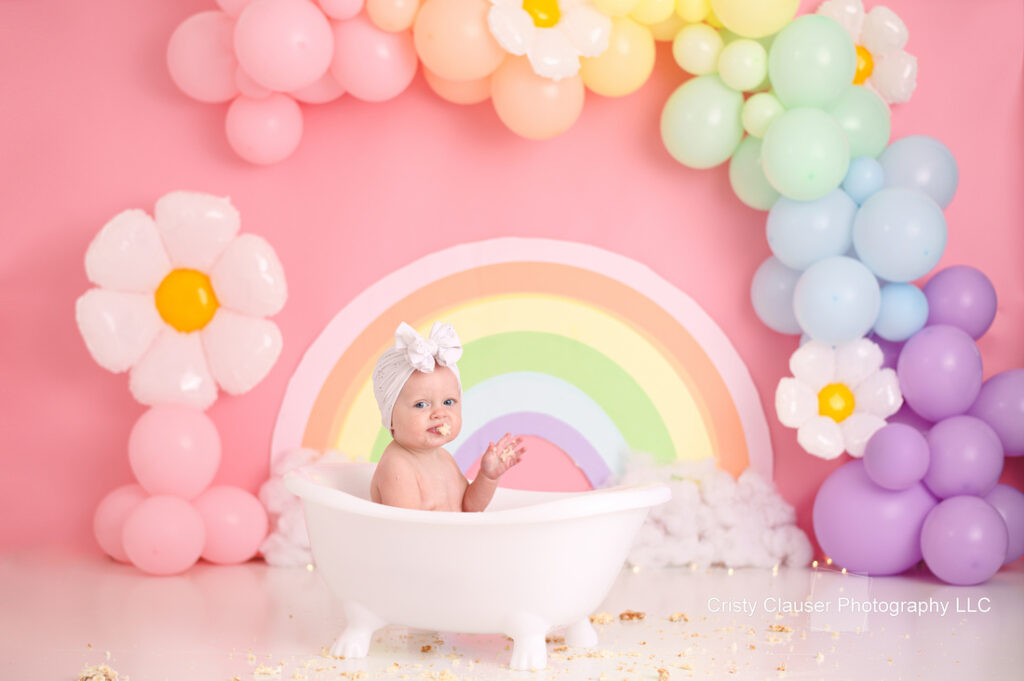 A baby in a white tub waves while surrounded by pastel balloons shaped like a rainbow and flowers. The background is a pink wall with a colorful rainbow design. The baby wears a white headband with a bow.