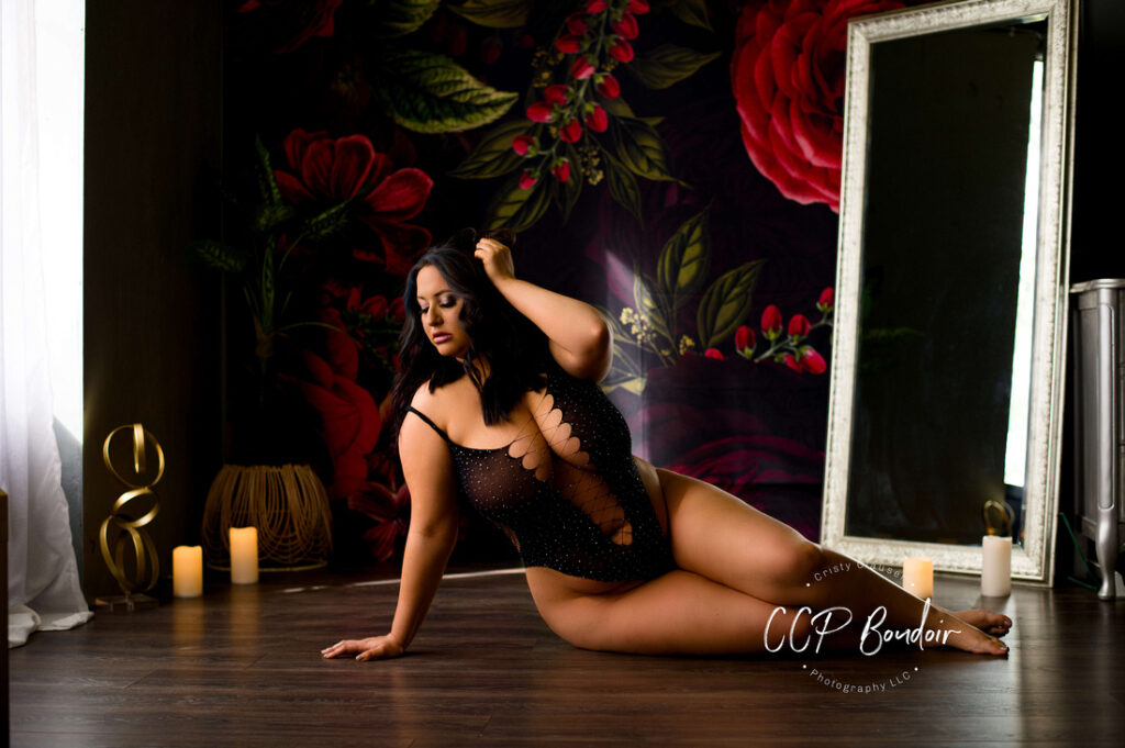 A woman in a sheer black bodysuit poses on a wooden floor beside lit candles, with a large mirror and dark floral mural in the background. The scene is softly lit, creating an intimate, elegant atmosphere. Cristy Clauser Photography