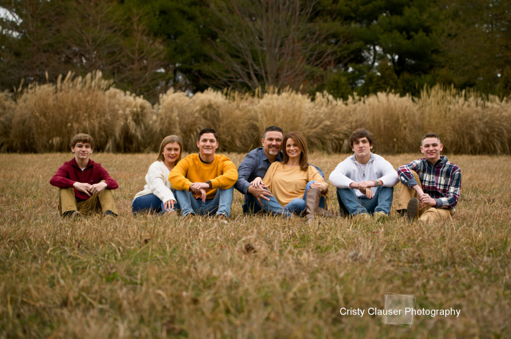 A family of seven sits in a grassy field, smiling at the camera. Tall brown grass and trees are in the background. The group is dressed in casual fall clothing. The image is credited to Cristy Clauser Photography. Cristy Clauser Photography