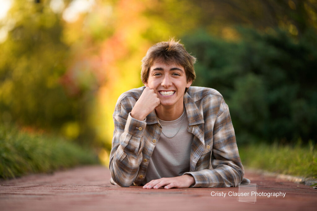 A young adult with brown hair smiles broadly while lying on a brick path. They wear a plaid shirt over a white top. The background is a blurred mix of green and autumn colors. Text on the image reads "Cristy Clauser Photography.