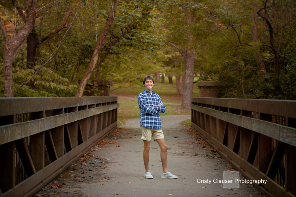 A person in a blue plaid shirt and khaki shorts stands confidently on a bridge in a wooded area. They are smiling and have their arms crossed. Trees with green leaves surround the path. Text on the image reads "Cristy Clauser Photography.