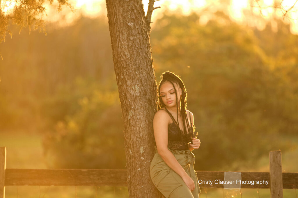 A woman with long braided hair leans against a tree at sunset, wearing a black lace top and olive green pants. The background features soft-focus greenery and warm golden light.