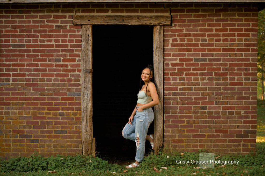 A person wearing a light green top and ripped jeans leans against the wooden frame of an open brick doorway, smiling. The sun casts a warm glow on the scene, highlighting the texture of the bricks and grass in the foreground.