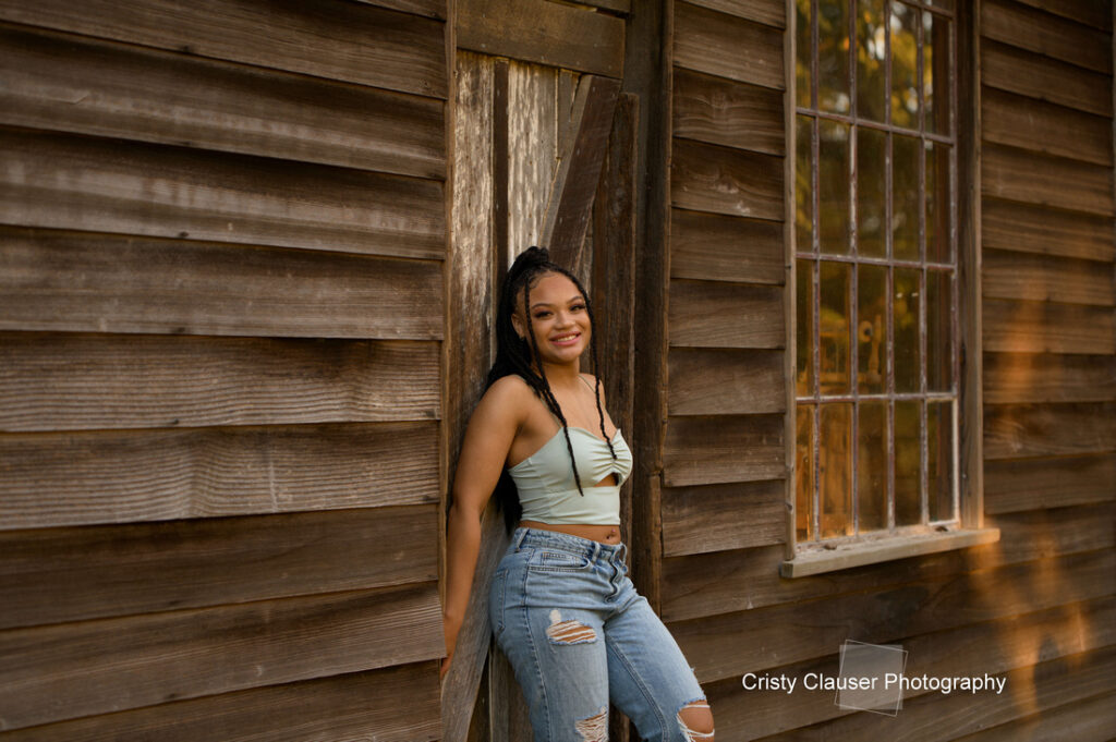 A woman with braided hair leans against a wooden wall next to a window. She is wearing a light-colored top and ripped jeans, smiling softly. Sunlight casts a warm glow on the scene. The photo is credited to Cristy Clauser Photography.