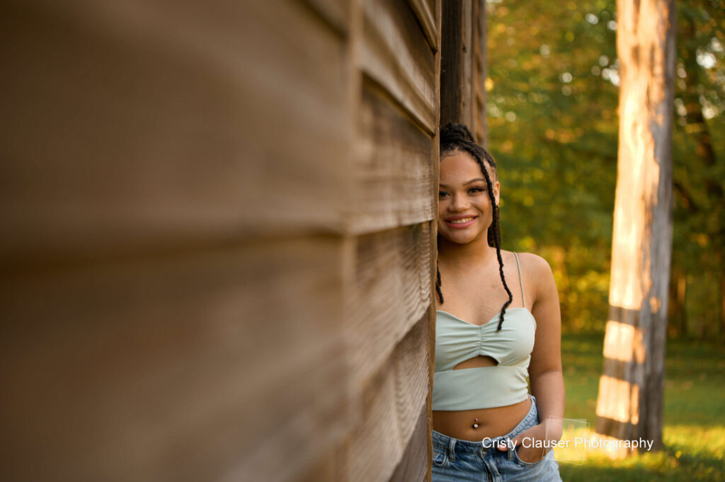 A young woman with braided hair and a pale green crop top leans against a wooden structure, smiling. She stands in a sunlit area with lush green foliage in the background. .