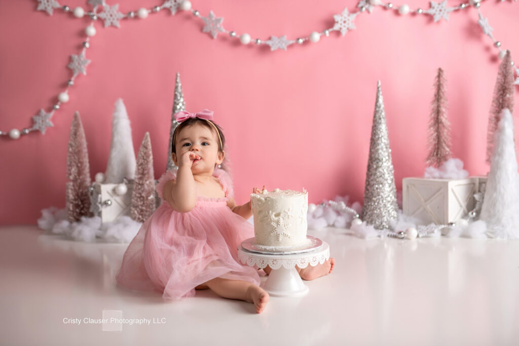 A baby in a pink dress sits on the floor, eating cake with her hand. The background is decorated with pink and white Christmas trees, garlands, and snowflakes on a pink wall. Cristy Clauser Photography