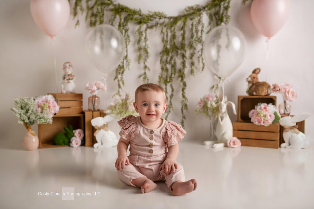A smiling baby sits on the floor in a pink outfit, surrounded by wooden crates, flowers, balloons, plush bunnies, and greenery hanging on the white wall, creating a soft, spring-themed setting. Cristy Clauser Photography