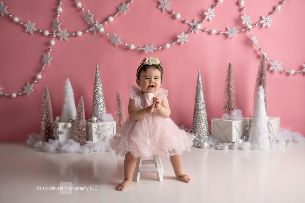 A baby girl in a pink dress and floral headband sits on a small white stool, surrounded by silver and white Christmas trees and ornaments, with a pink backdrop decorated with snowflakes and garlands. Cristy Clauser Photography
