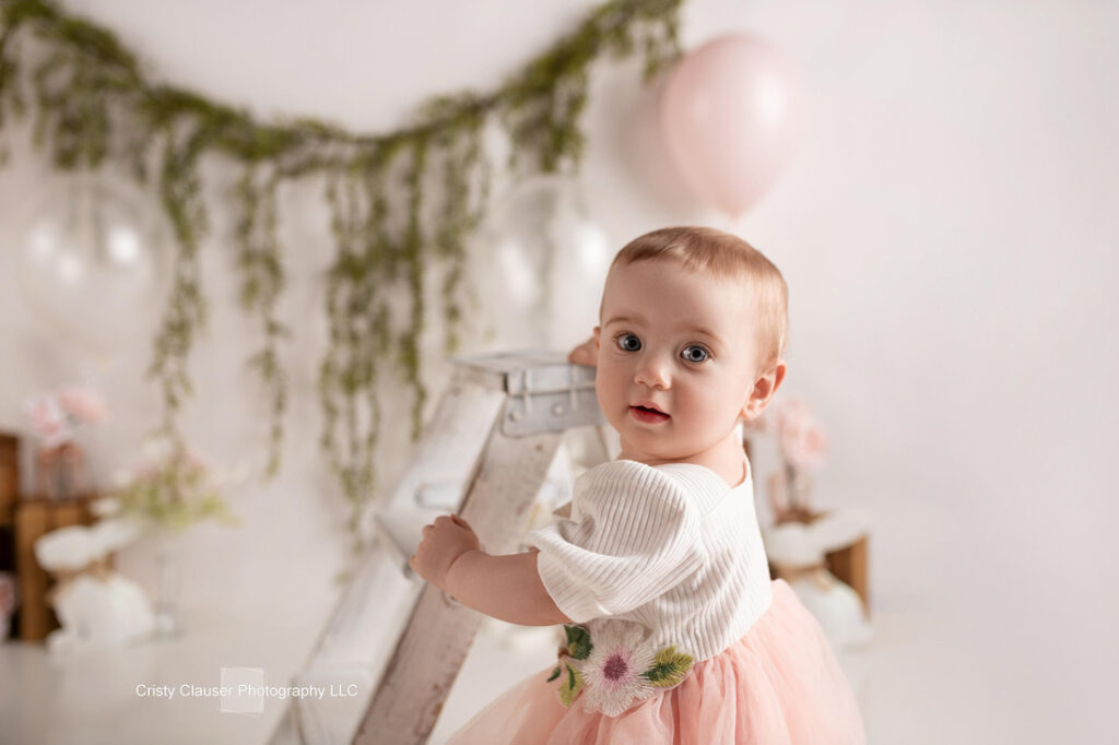A baby in a white top and pink tulle skirt stands holding a white ladder, looking at the camera. The background features green garland, pink and white balloons, and soft, pastel decorations. Cristy Clauser Photography