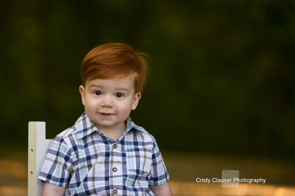 A young child with red hair and a plaid shirt sits on a white chair outdoors, smiling gently with a blurred green background behind. The photo is credited to Cristy Clauser Photography. Cristy Clauser Photography