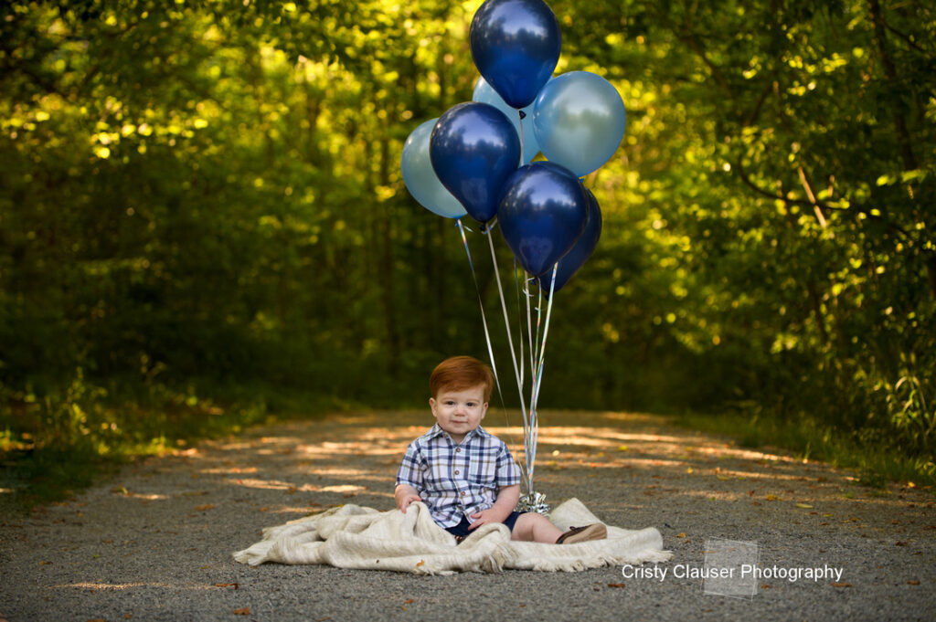 A young boy with red hair sits on a blanket on a forest path, holding several blue balloons. Sunlight filters through the green trees in the background. "Cristy Clauser Photography" is written at the bottom right. Cristy Clauser Photography