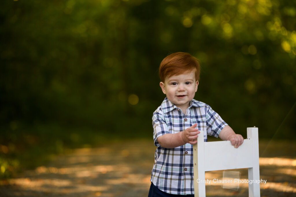 A young child with red hair, wearing a plaid shirt, stands outdoors holding the back of a white wooden chair on a sunlit path surrounded by greenery. Cristy Clauser Photography