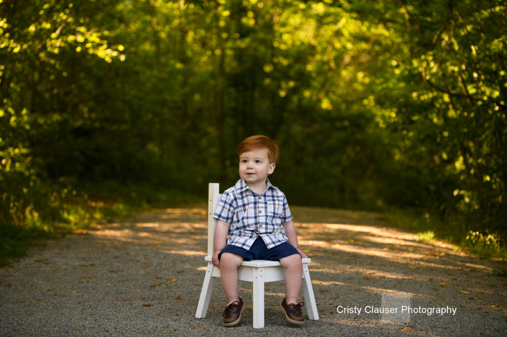 A young boy with red hair sits on a small white chair in the middle of a sunlit forest path, wearing a plaid shirt, blue shorts, and brown shoes. Lush green trees surround the scene. Cristy Clauser Photography