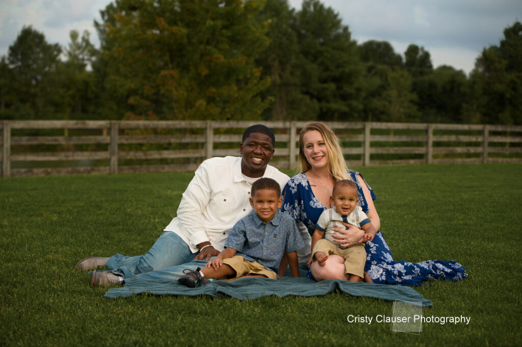 A smiling family of four sits on a blanket in a grassy field. The parents sit behind their two young sons. Trees and a wooden fence are in the background. The mother wears a blue dress; the father and children wear light-colored clothes. Cristy Clauser Photography