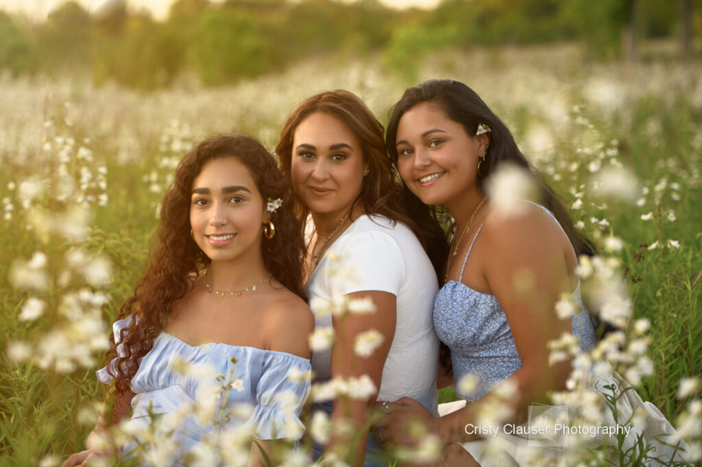 Three young women sit closely together in a field of white wildflowers at sunset, smiling at the camera. They wear light blue and white outfits, and flowers are tucked behind their ears. The scene is warm and serene. Cristy Clauser Photography