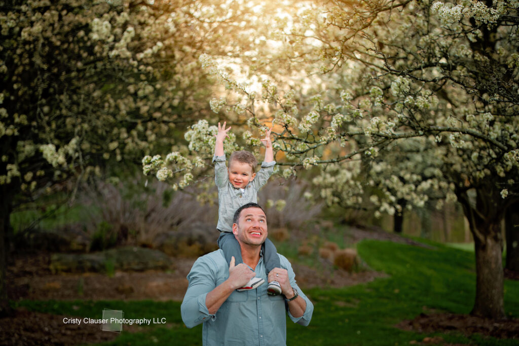 A man gives a smiling young child a shoulder ride under blooming trees, with sunlight filtering through the branches in a green, outdoor park. Both look happy, and the child reaches up toward the flowers. Cristy Clauser Photography