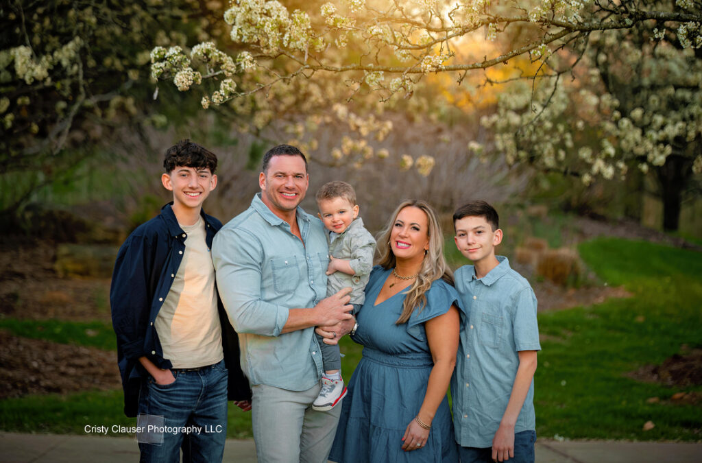 A smiling family of five poses outdoors under blooming trees at sunset. The parents stand in the center with their young child, flanked by two older boys. All wear coordinated blue and gray outfits. Cristy Clauser Photography