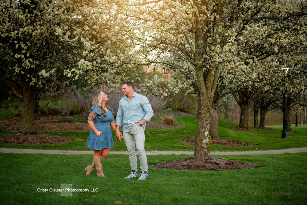 A couple walks hand in hand on green grass beneath blooming trees in a sunlit park, smiling at each other. The woman wears a blue dress and boots; the man wears a light shirt and jeans. Cristy Clauser Photography