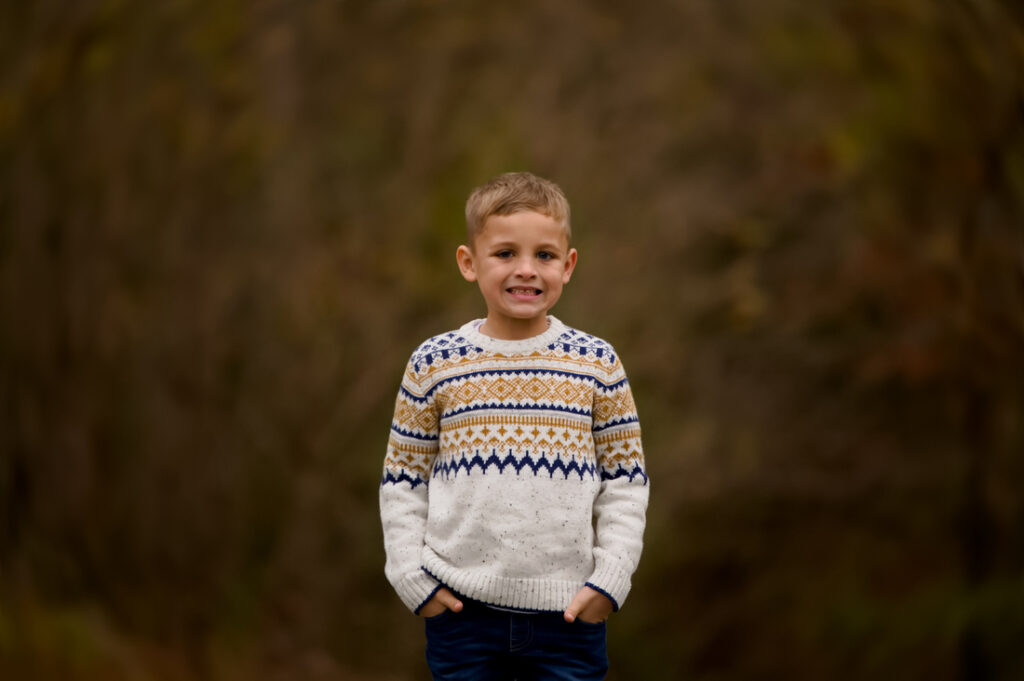 A young boy stands outdoors, smiling with his hands in his pockets. He is wearing a white sweater with a colorful geometric pattern, and the background is blurred with earthy tones. Cristy Clauser Photography