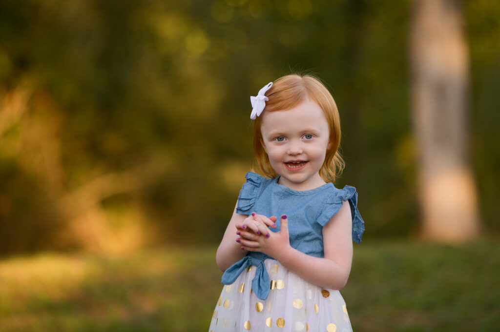 A young girl with red hair, wearing a blue and white dress with gold polka dots and a white bow, stands outdoors smiling with hands clasped. The background is green and softly blurred. Cristy Clauser Photography