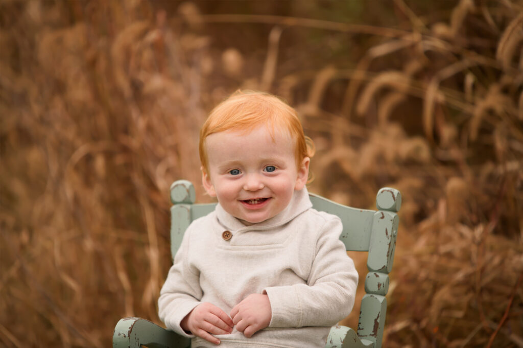 A smiling young child with red hair sits on a pale green wooden chair outdoors, surrounded by tall, golden-brown grasses. The child is wearing a light beige sweater. Cristy Clauser Photography