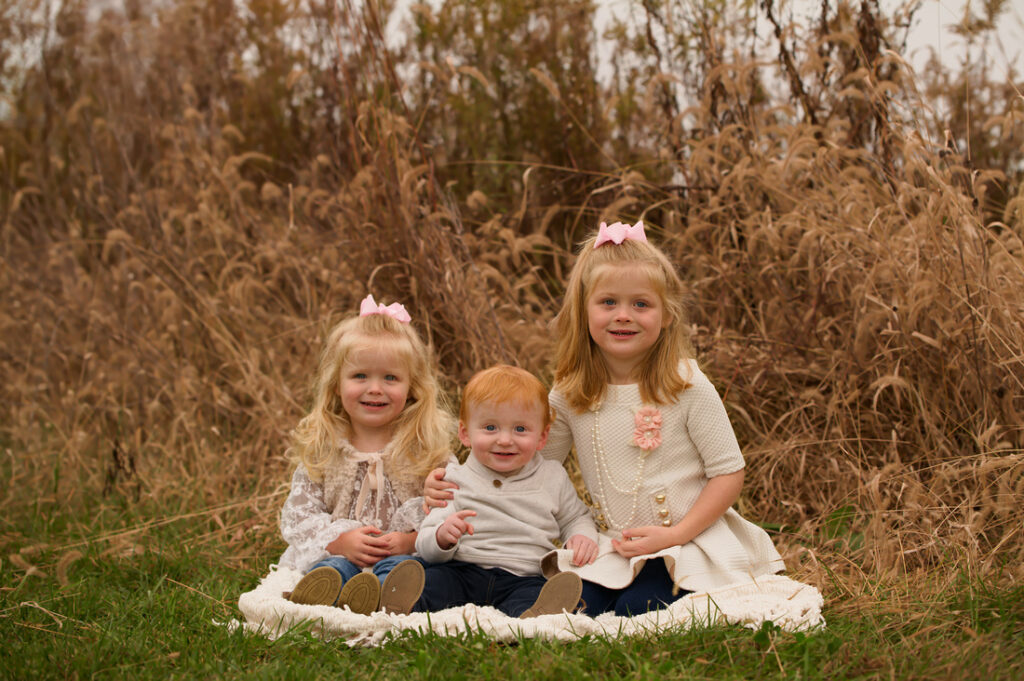 Three young children sit together on a blanket outdoors in front of tall brown grass. Two blonde girls in light dresses and pink bows flank a red-haired toddler boy in gray. All three are smiling at the camera. Cristy Clauser Photography
