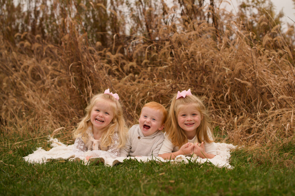 Three young children, two girls with blonde hair and pink bows and a red-haired baby boy, lie on a white blanket in the grass, smiling in front of tall, dried brown grasses. Cristy Clauser Photography