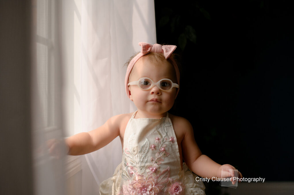 A young toddler wearing glasses, a light dress with floral embroidery, and a pink bow headband stands by a window with sheer curtains, softly lit by natural light. Cristy Clauser Photography