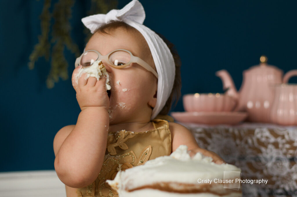 A baby with glasses and a white headband eats cake with her hand, getting frosting on her face. Pink teapots and teacups are in the background on a lace-covered table. Cristy Clauser Photography