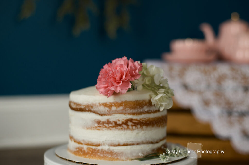 A small, semi-naked cake decorated with pink and white flowers sits on a white stand. In the blurred background, there are pink teapots and teacups on a lace-covered table. Cristy Clauser Photography