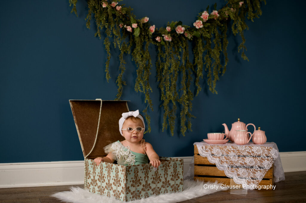 A baby wearing glasses, a white bow, and a lace outfit sits in a decorative trunk on a fur rug. Behind is a dark blue wall with a floral garland, and a table with pink tea set and lace cloth. Cristy Clauser Photography