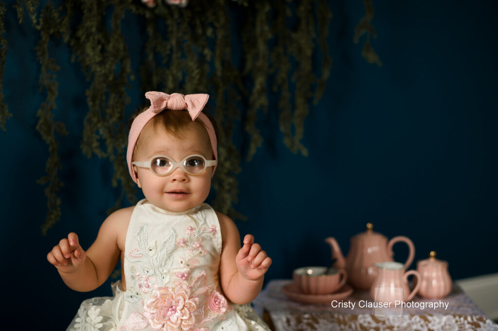 A young child wearing glasses, a pink headband, and a white floral dress sits in front of a dark blue background with greenery hanging above. A pink tea set is arranged on a table beside her. Cristy Clauser Photography