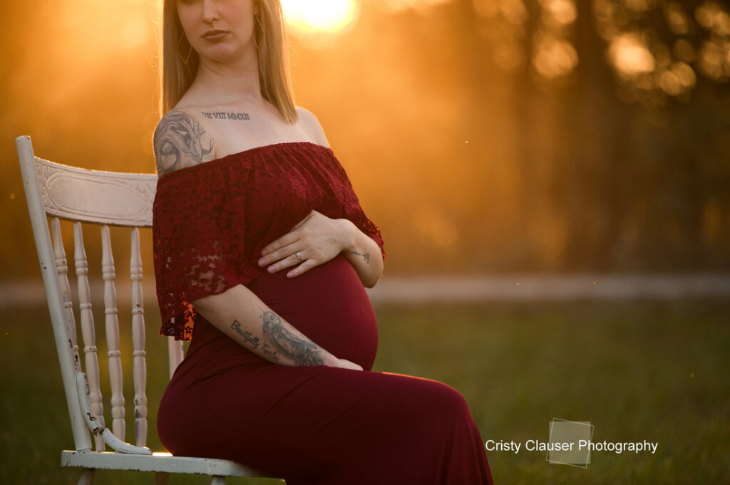 A pregnant woman in a red dress sits on a white chair outdoors at sunset, gently cradling her belly. Sunlight creates a warm, golden glow around her. "Cristy Clauser Photography" is noted in the bottom right corner. Cristy Clauser Photography