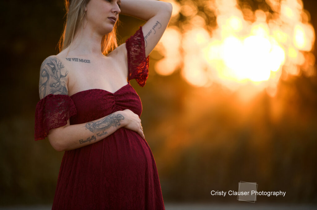 A pregnant woman in a burgundy off-shoulder dress cradles her belly and rests one arm on her head, standing outdoors with sunlight glowing in the background. Tattoos are visible on her arms and chest. Cristy Clauser Photography