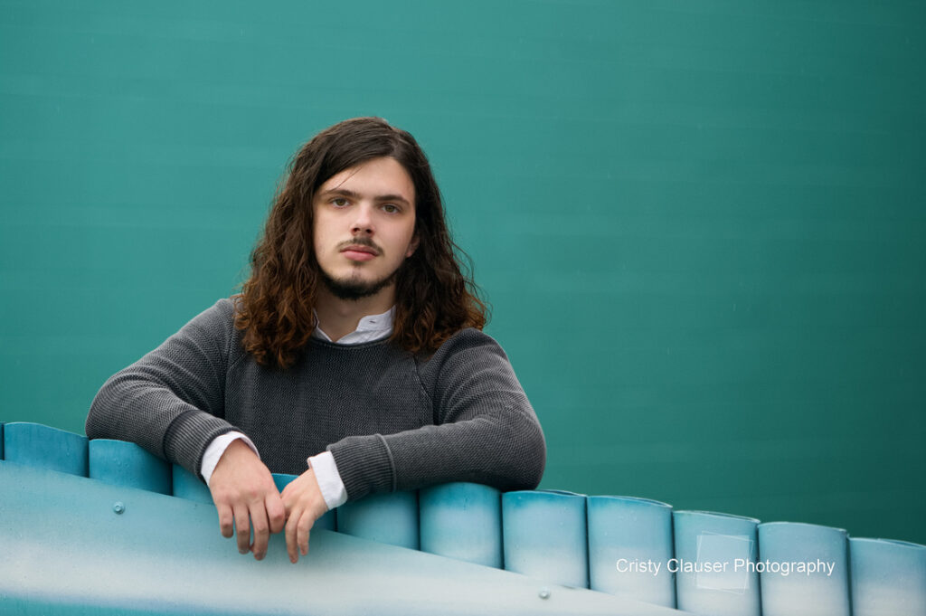 Person with long hair and beard, wearing a gray sweater over a white shirt, leaning on a blue fence. The background is a solid teal color.
