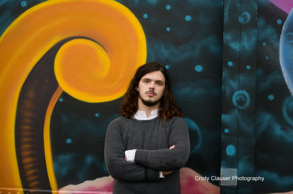 A person with long hair and a gray sweater stands with arms crossed in front of a vibrant, swirling mural. The background features bold, colorful patterns in yellow, orange, and blues. The words "Cristy Clauser Photography" are in the corner.