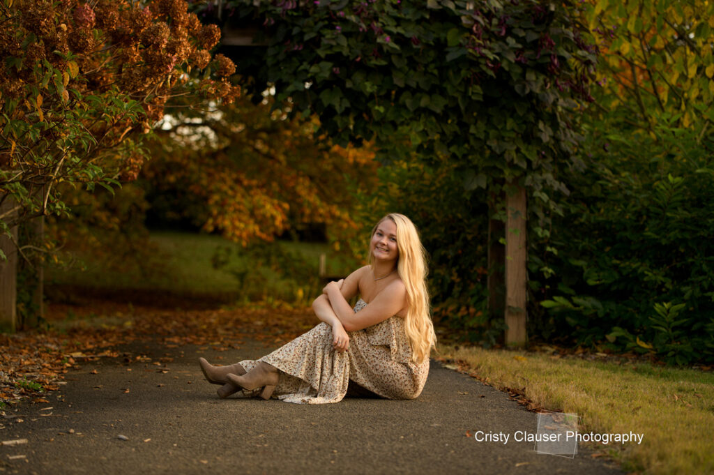 A woman with long blonde hair, dressed in a floral dress and boots, sits smiling on a path. She is surrounded by lush greenery and autumnal leaves in a peaceful outdoor setting.
