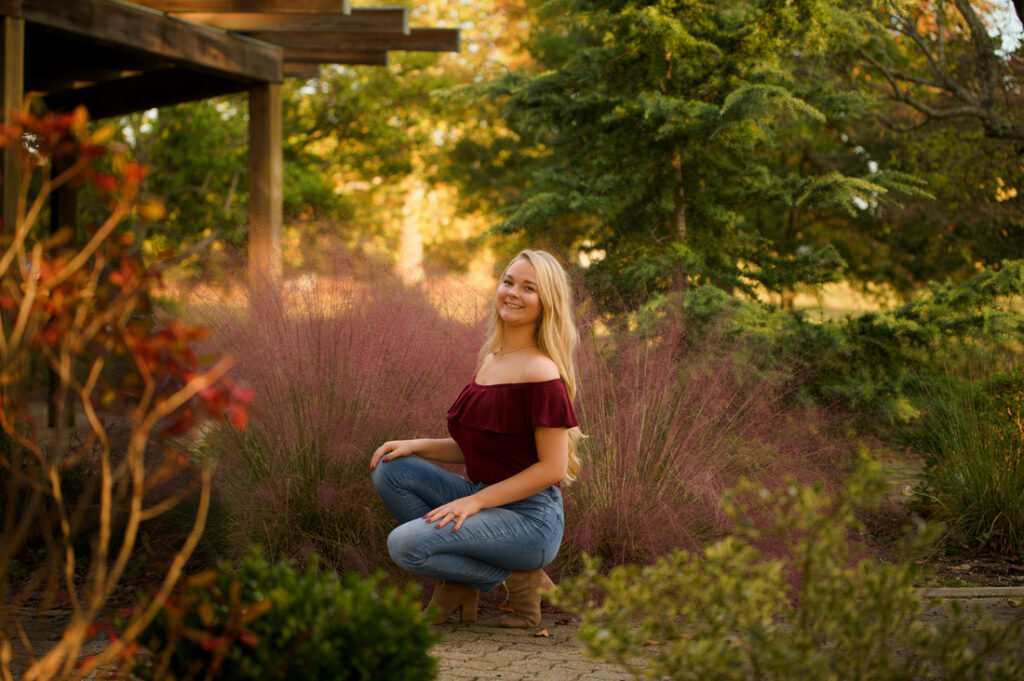 A person squats in a garden, wearing a burgundy off-shoulder top and blue jeans, surrounded by green foliage and pink ornamental grass. The setting is serene and sunlit, with blurred autumn colors in the background.