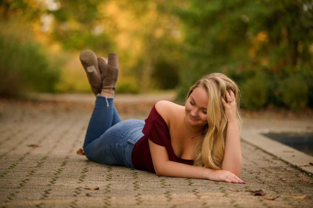 Woman lying on her stomach on a paved pathway, smiling and touching her hair. She is wearing a burgundy off-shoulder top, blue jeans, and brown ankle boots. The background features blurred greenery, suggesting a park or garden setting.