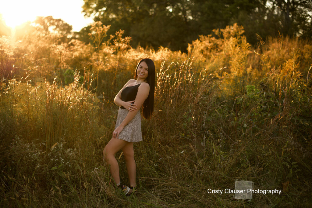 A woman with long hair stands smiling in a sunlit field with tall grass and wildflowers. She wears a black top and patterned shorts, surrounded by golden light. The sky is bright, and trees are visible in the background.