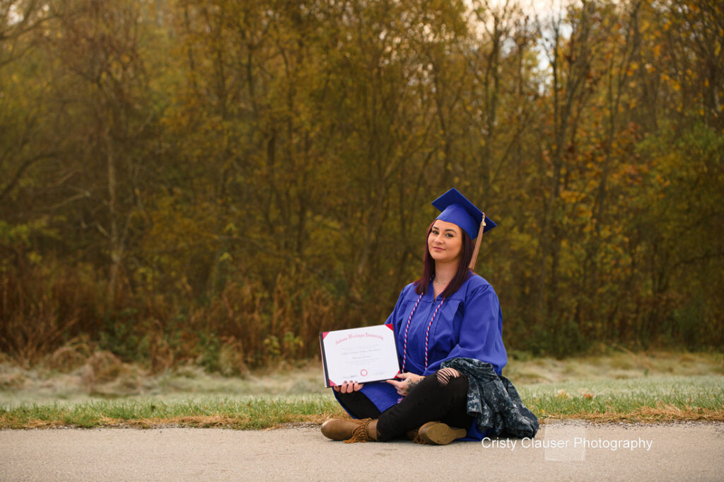 A graduate in a blue cap and gown sits on the grass holding a framed certificate. The background shows trees and a misty field.