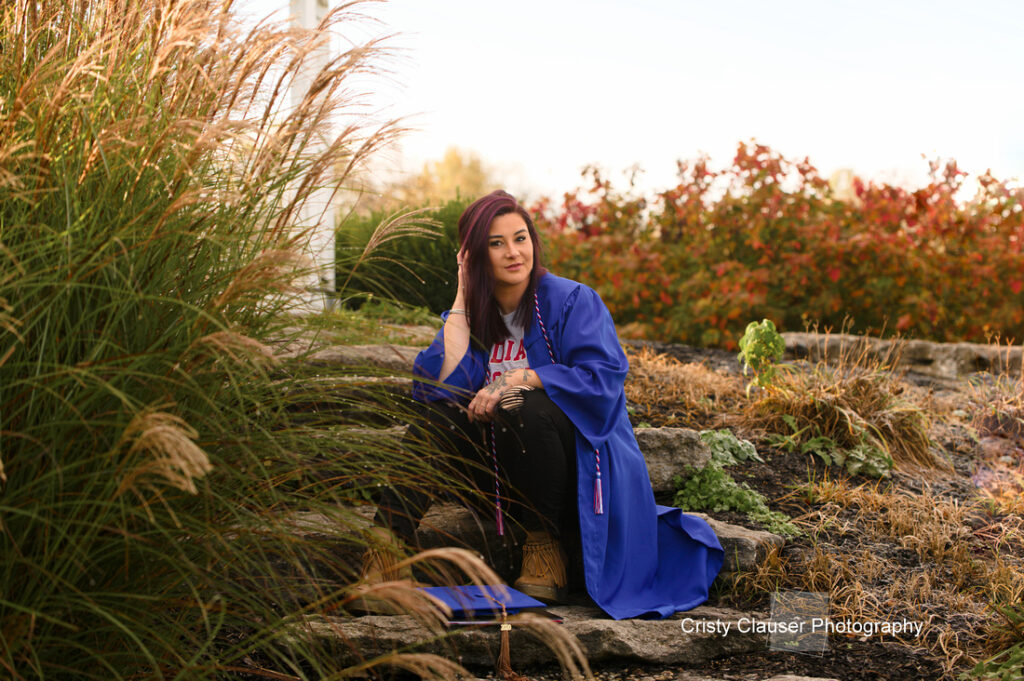 A person in a blue graduation gown sits outdoors on stone steps, surrounded by tall grasses and red foliage. They are smiling slightly, with a cap on their lap. The sky is clear, indicating a sunny day.